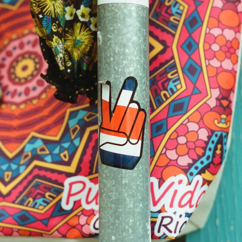 A sticker of a hand making a peace sign in red, white, and blue colors is on a metal pole. In the background, there's a colorful, patterned object with text partially reading "Pura Vida.