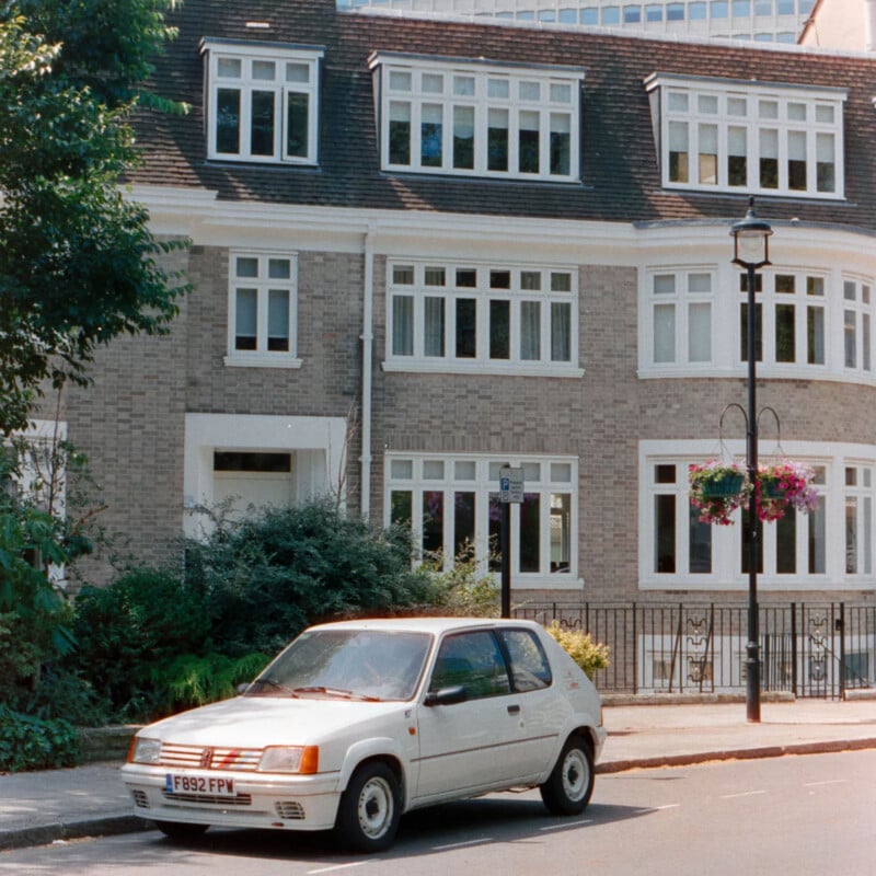 A white hatchback car is parked on a quiet street in front of a three-story brick building with many white-framed windows. There are trees, greenery, and hanging flower baskets nearby.