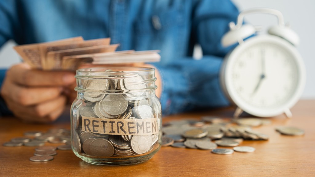 A person putting money into a glass jar labeled "RETIREMENT," with more money and an alarm clock in the background.