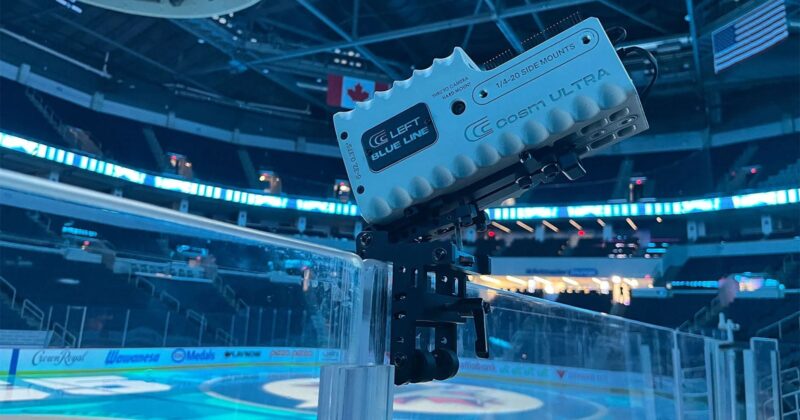 A close-up of a video camera mounted on the glass boards of an indoor ice hockey rink, with empty seats and Canadian and American flags visible in the arena background.