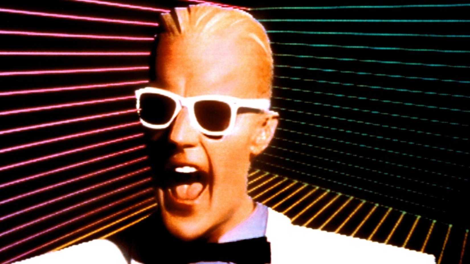 Max Headroom smiling in front of an 1980s style backdrop.
