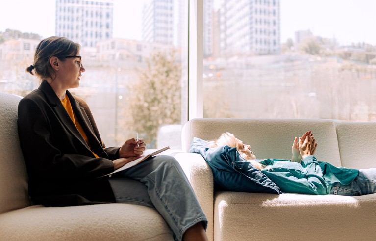 A psychologist with a pen and pad talks with a patient reclining on a sofa during a therapy session.