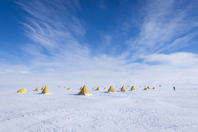 A vast snowy landscape dotted with a cluster of yellow tents and a lone figure.