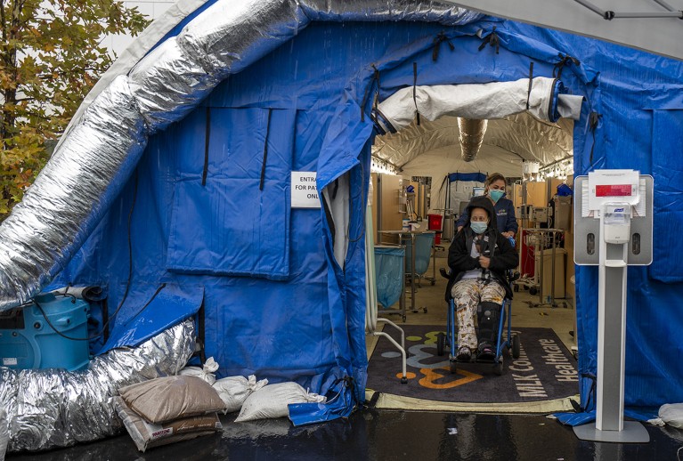 Two people wearing face masks in a blue tent, from which emerge silver-coloured pipes.