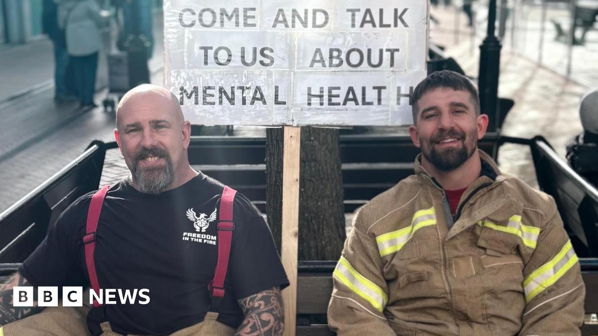 Firefighters Lee Ralph and Warren Shepherd dressed in uniform, sat on a bench. Behind them is a sign that says ' Come and talk to us about mental health'.