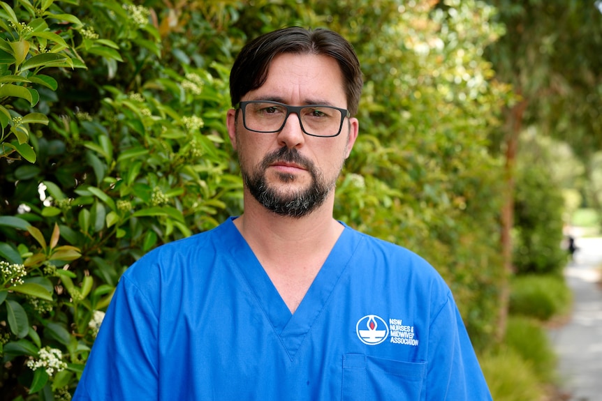 A bearded man in a blue nursing scrubs looks unsmiling at the camera.
