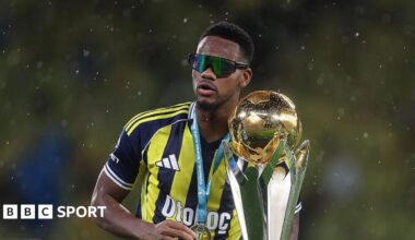 Jhon Duran in Fenerbahce's yellow and navy blue vertical-striped kit, with a large trophy, while wearing sunglasses