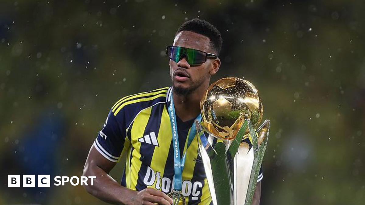 Jhon Duran in Fenerbahce's yellow and navy blue vertical-striped kit, with a large trophy, while wearing sunglasses