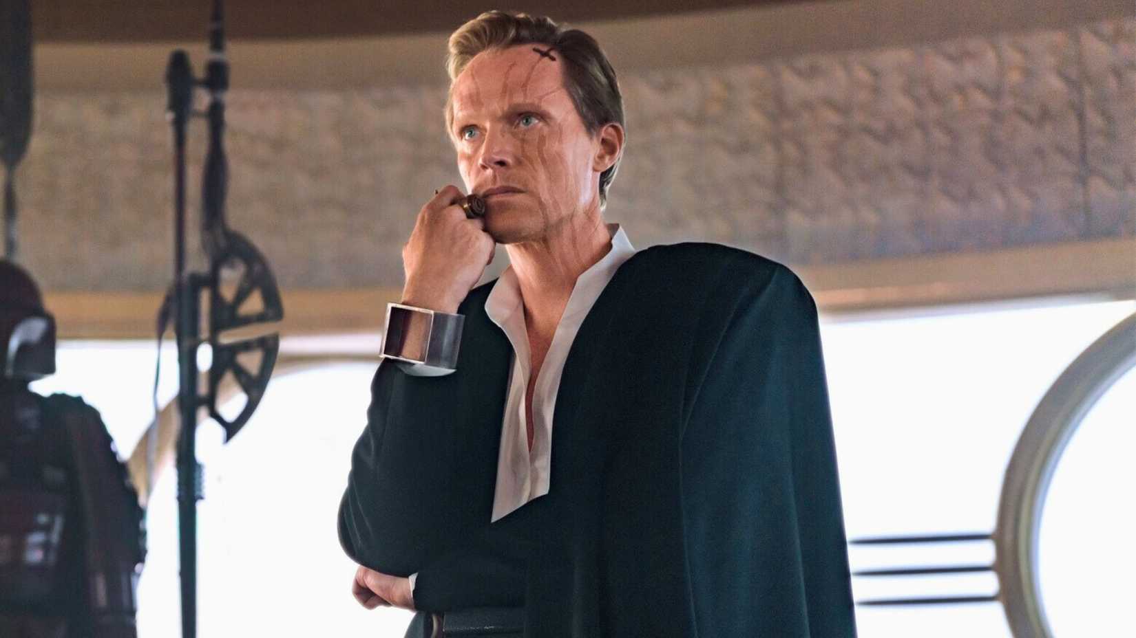 An image of Paul Bettany as Dryden Vos in the 2018 film, Solo: A Star Wars Story. Dryden is dressed in a black and white suit. He has a hand pressed to his face, and looks menacing.