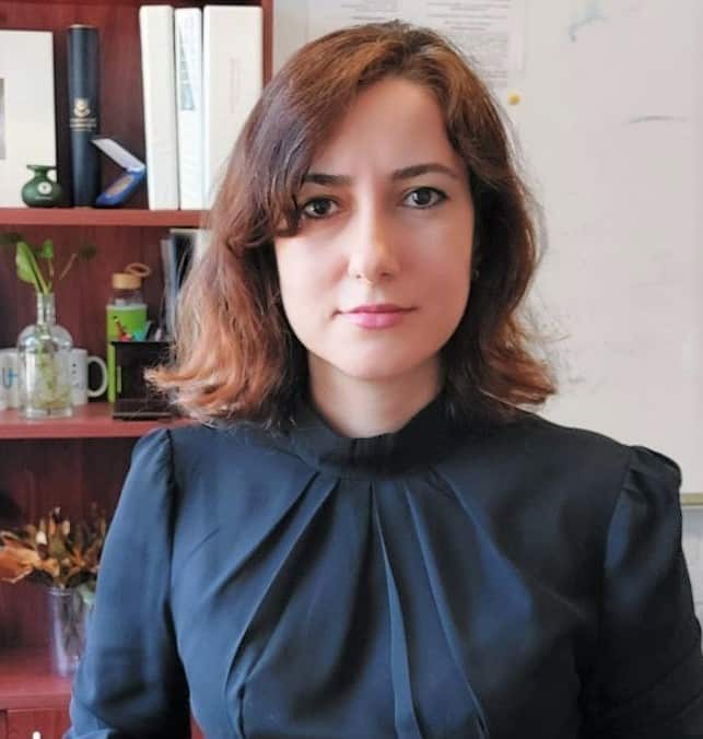 A woman with shoulder length brown hair and wearing a blue shirt sits in front of a book case.
