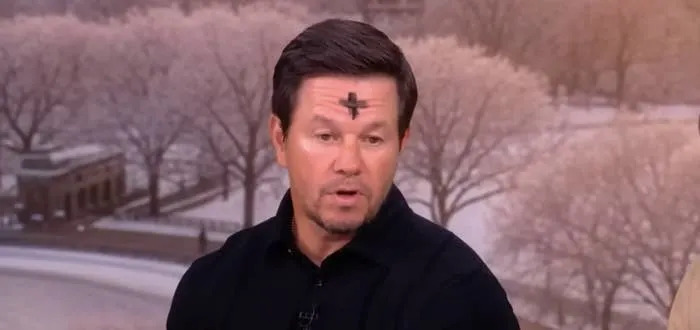 Mark Wahlberg with a cross on forehead speaking during an interview, with a wintry park scenery in the background