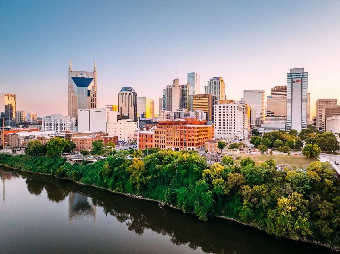 A view of downtown Nashville, Tennessee at sunset, featuring modern skyscrapers, the iconic AT&T Building, and lush green trees along the river in the foreground.