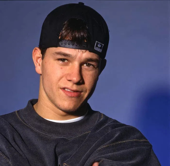 Mark Wahlberg wearing a backwards cap and sweatshirt, facing the camera with arms crossed in a neutral pose