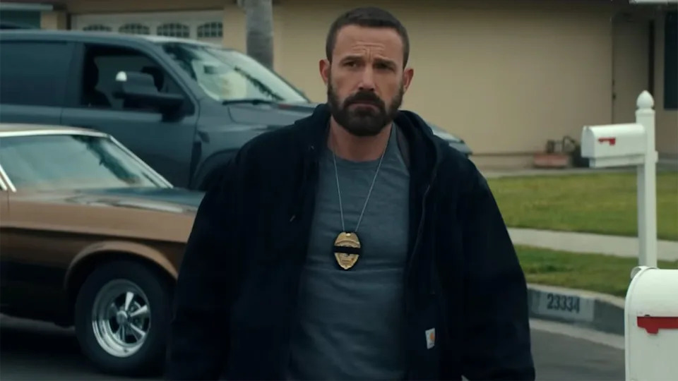  Ben Affleck in a tee and jacket and wearing a cop badge outside of a home in The Rip. 