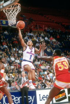Muggsy Bogues in mid-air, arm extended to the net with basketball in hand, players of the competing team surrounding him