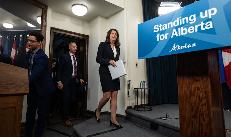 A dark-haired woman walks towards a podium and a sign that reads Standing up for Alberta.