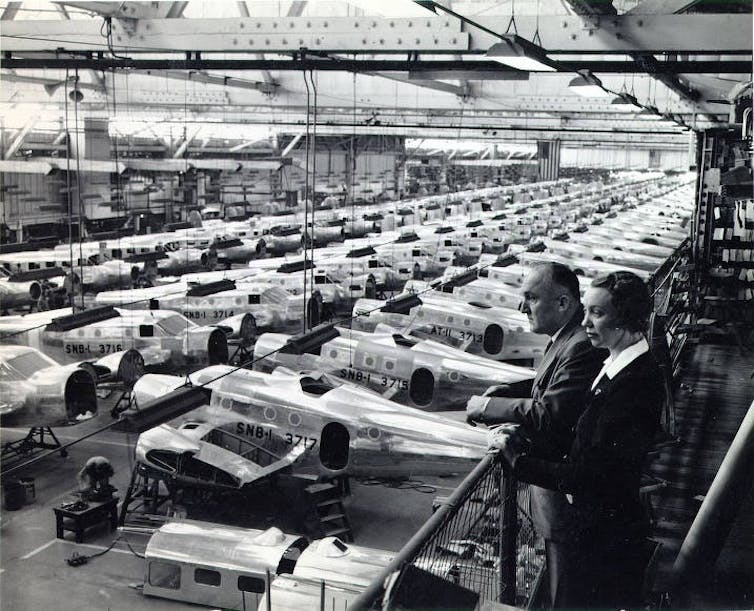 Two people look over a production line with dozens of military aircraft in a large building.