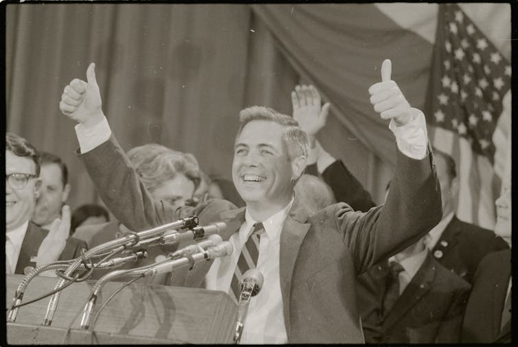 A man in a sports jacket and tie, gives thumbs up as he stands behind a lectern featuring microphones.