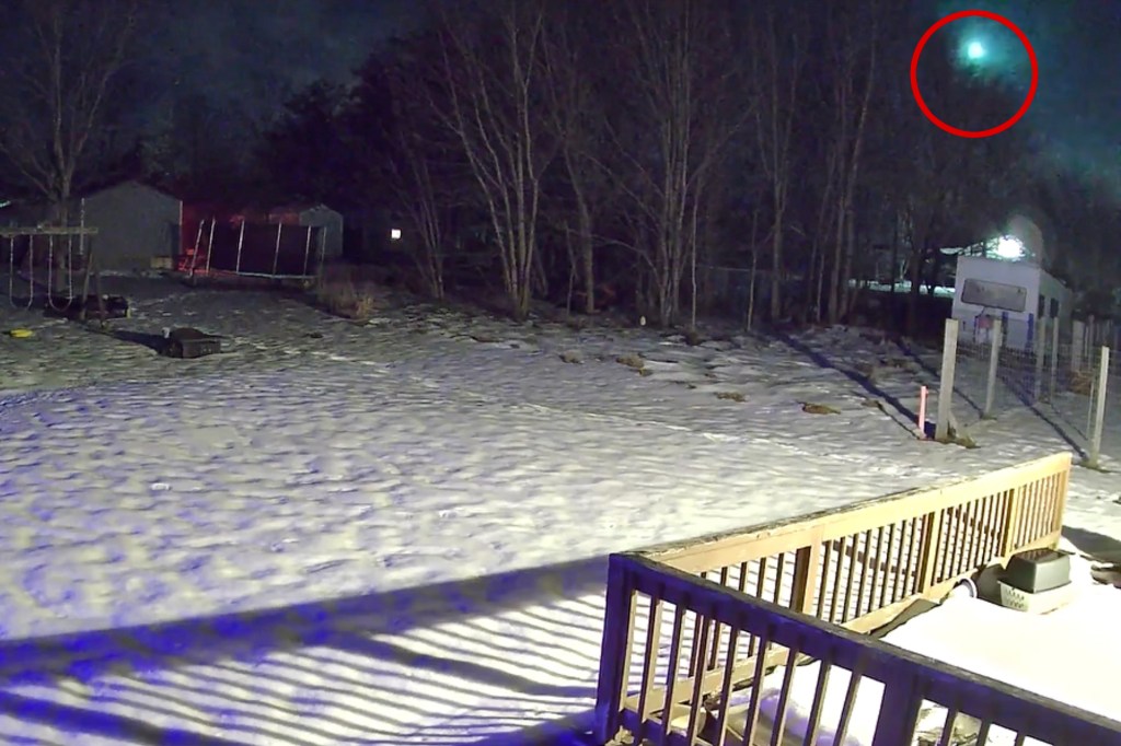 A fireball streaks through the night sky, visible through trees in a snow-covered yard.