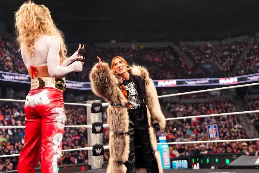 Becky Lynch stands facing the viewer in a wrestling ring, teeth bared, pointing and glaring at Maxxine Dupri, whose back is to the camera. Lynch is wearing a shaggy tan fur coat and holding a microphone down by her side; Dupri, holding a microphone of her own, is in red pants with white graffiti designs and a white shirt, and she wears a thick white leather championship belt with gold trimmings.
