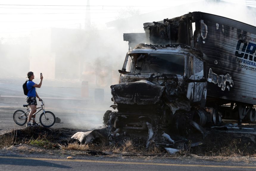 A man riding a bicycle takes a photo of a burned truck, allegedly set on fire by organised crime groups on a highway near Acatlan de Juarez, Jalisco state, Mexico, on February 22, 2026.