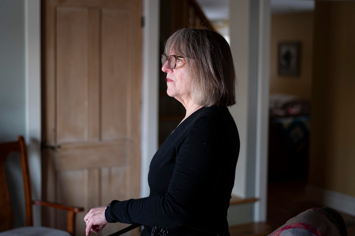 A woman with shoulder-length gray hair and glasses stands indoors in profile, wearing a black top and looking forward.