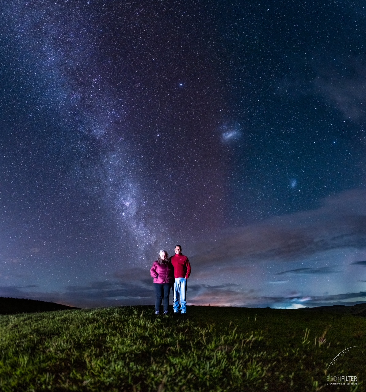 two people stand smiling at the camera against the background of the milky way stretching into the sky and a distinct red glow behind them, a diffuse glow filling the sky.