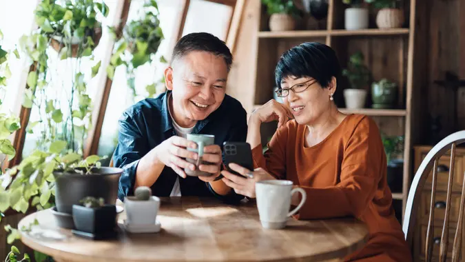 A happy retired couple smiles and drinks coffee while sitting at their kitchen table with plants next to them