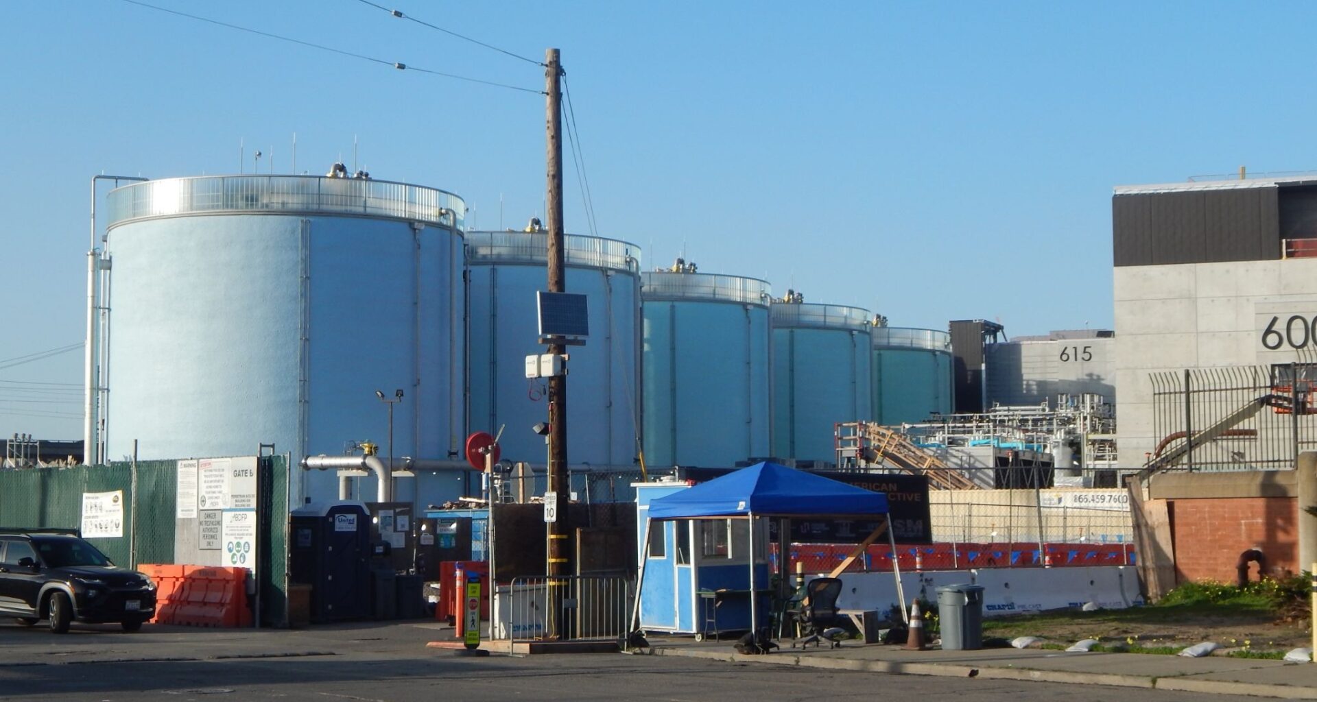 Large cylindrical industrial storage tanks are situated behind a gate and a small blue security booth, with various industrial equipment and buildings visible nearby.