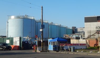 Large cylindrical industrial storage tanks are situated behind a gate and a small blue security booth, with various industrial equipment and buildings visible nearby.