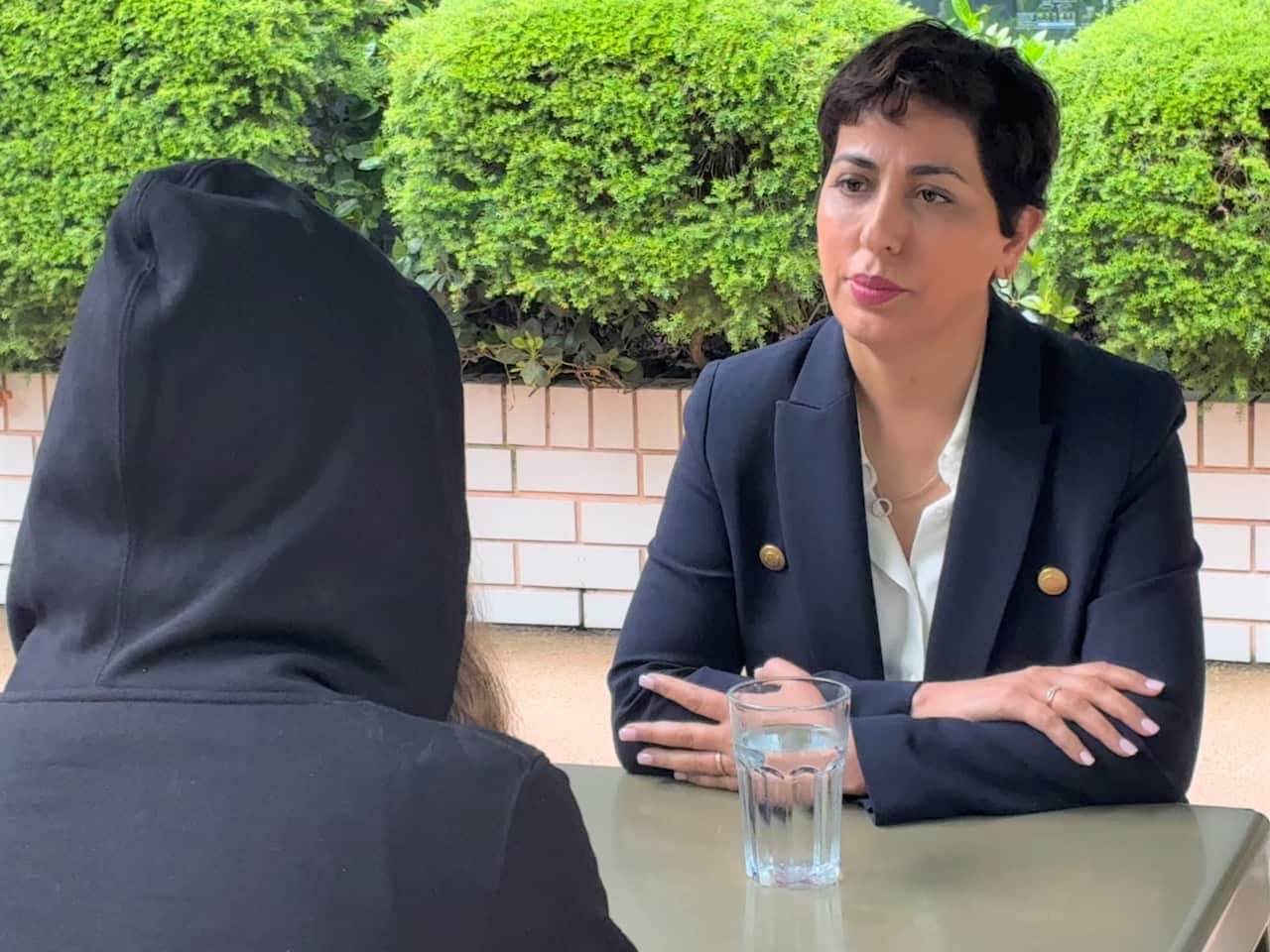 A woman wearing a blue blazer sits at a table with another person wearing a black hoodie.