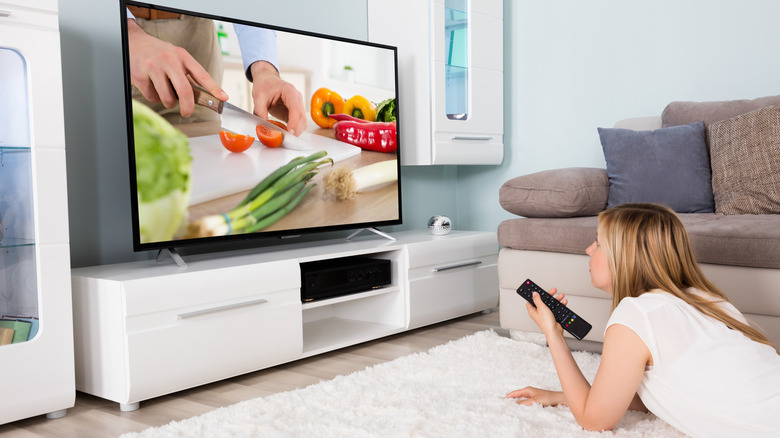 A woman lying on a living room floor, watching a cooking show on TV