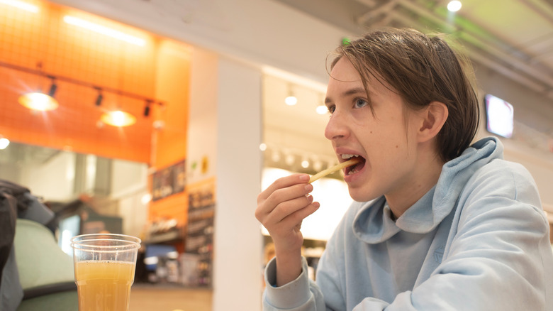 A person is seen eating French fries in a generic food court setting.