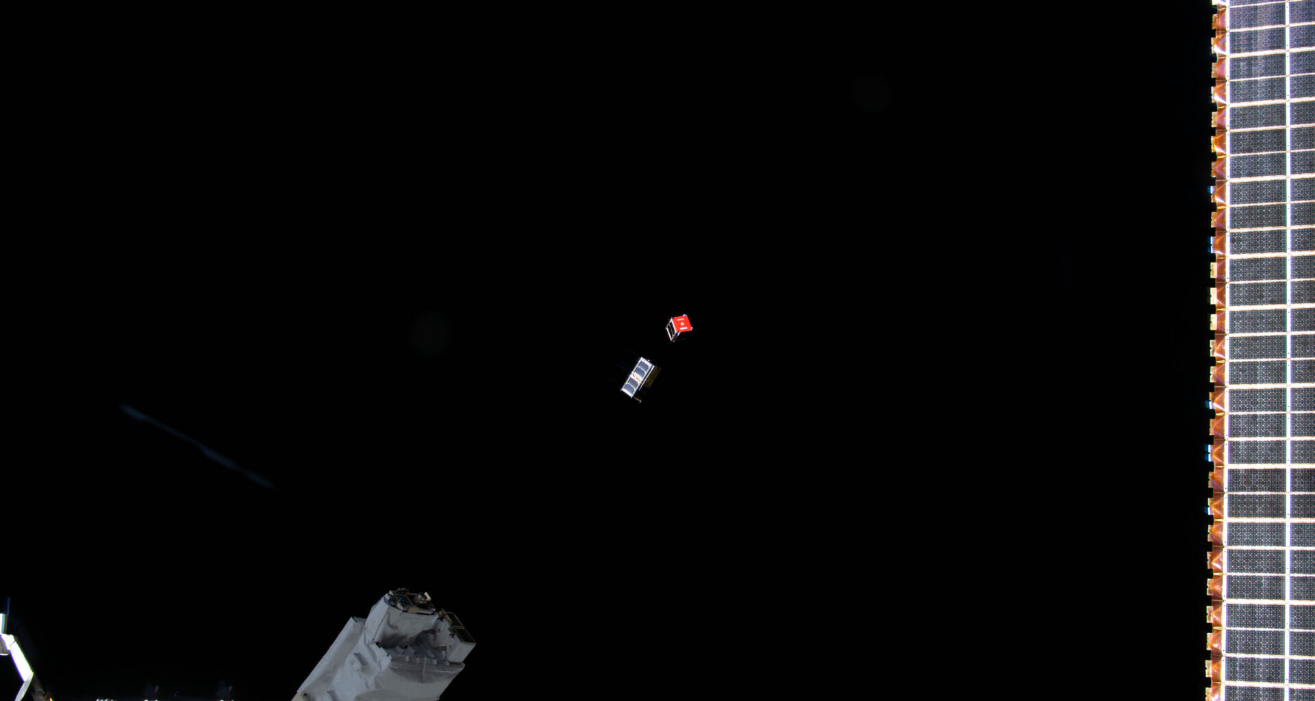 Two CubeSats, one silver and one with a red face, are ejected from the International Space Station (bottom left) into the blackness of space.