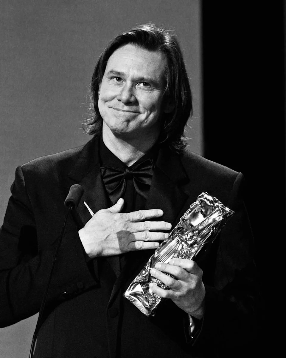 Jim Carrey holding an honorary César award while speaking on stage, wearing a black tuxedo and bow tie.