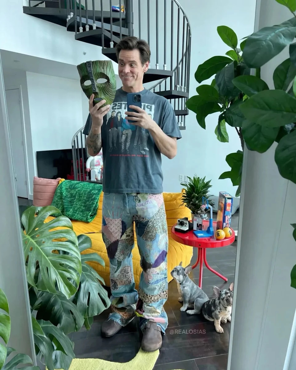 Jim Carrey taking a mirror selfie while holding a green carved mask resembling the one from the film The Mask, with two small dogs sitting nearby.