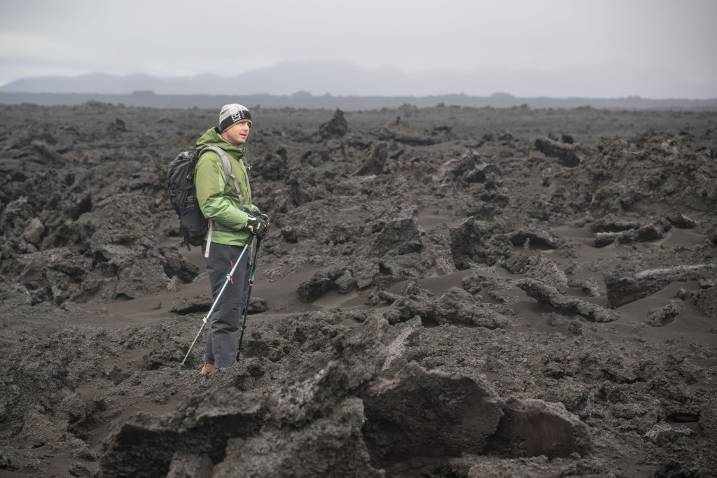 Reid Wiseman in a green jacket and stocking cap hikes across a Moon-like grey landscape in Iceland while participating in Artemis II Crew Geology Training in Iceland.