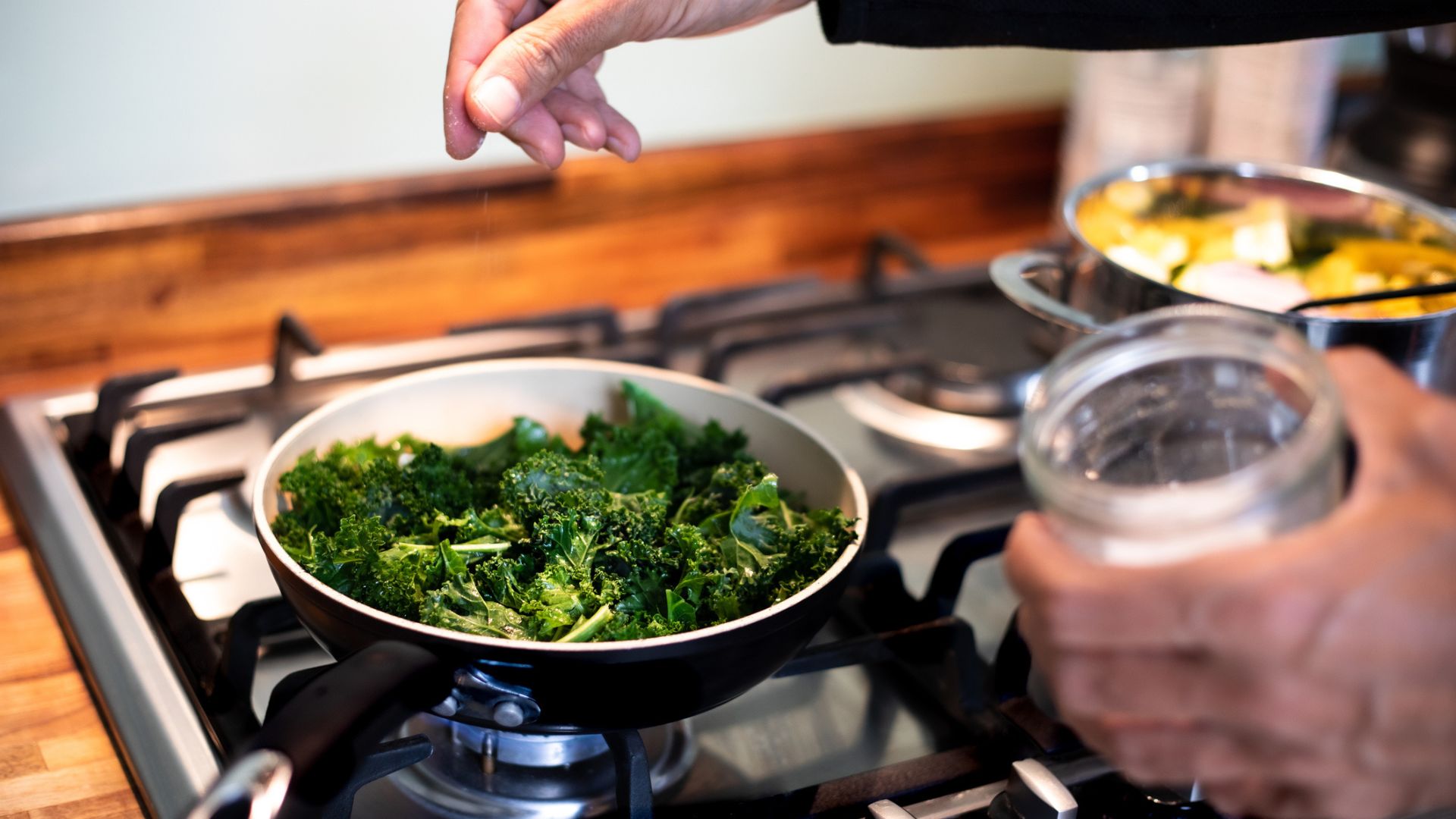 man's hand sprinkles herbs into a pan of cooking kale