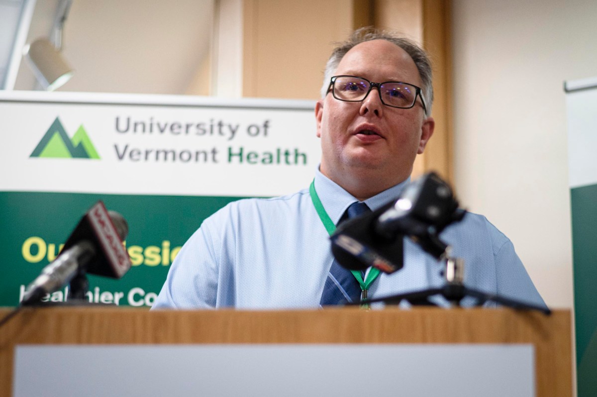 A man in a shirt and tie speaks at a podium with microphones, in front of a University of Vermont Health sign.