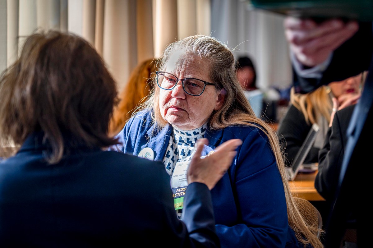 Two women sit indoors, engaged in a serious conversation; one woman listens intently, wearing glasses and a badge, while another gestures with her hand.