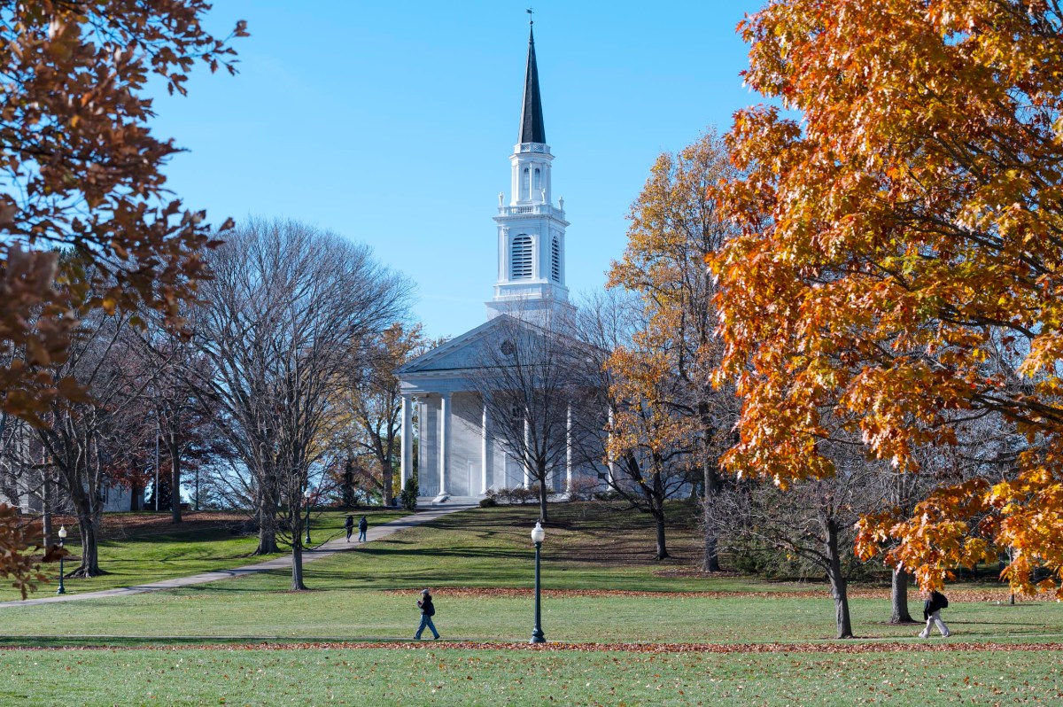 A white church with a steeple is surrounded by autumn trees and a green lawn, with a few people walking across the campus on a clear day.