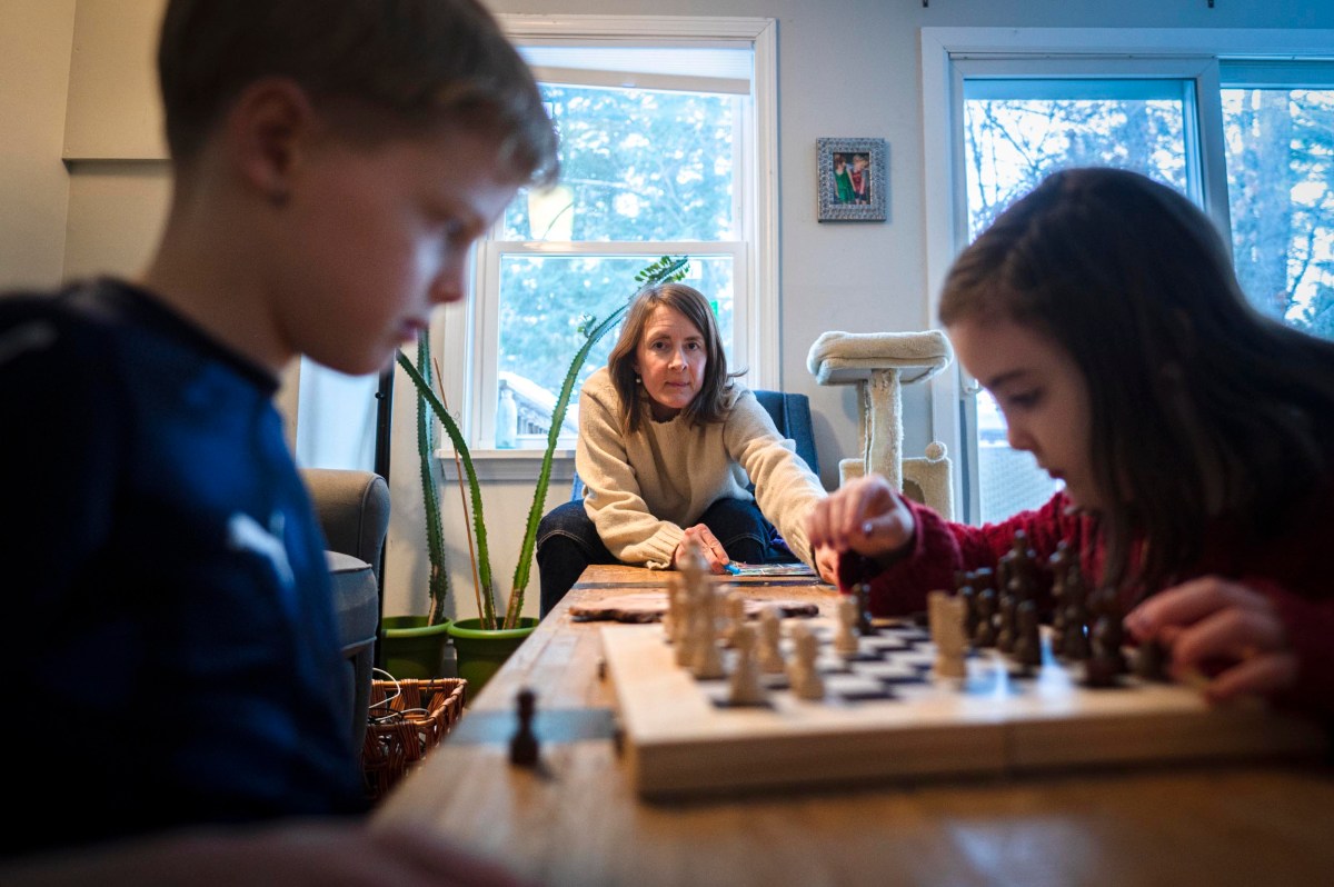 A woman watches two children play chess at a wooden table in a bright room with large windows and houseplants.