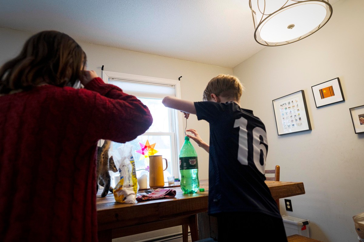 Two children stand at a wooden table with snacks and a green soda bottle in a well-lit room decorated with framed pictures and a hanging light fixture.