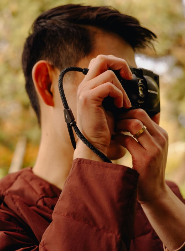 A person wearing a maroon jacket holds a black camera up to their face, preparing to take a photo outdoors. They are wearing a gold ring on their left hand. The background is softly blurred with greenery.