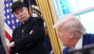 Elon Musk stands with his arms crossed, wearing a DOGE cap, in the Oval Office, near President Trump who sits at his desk while speaking.
