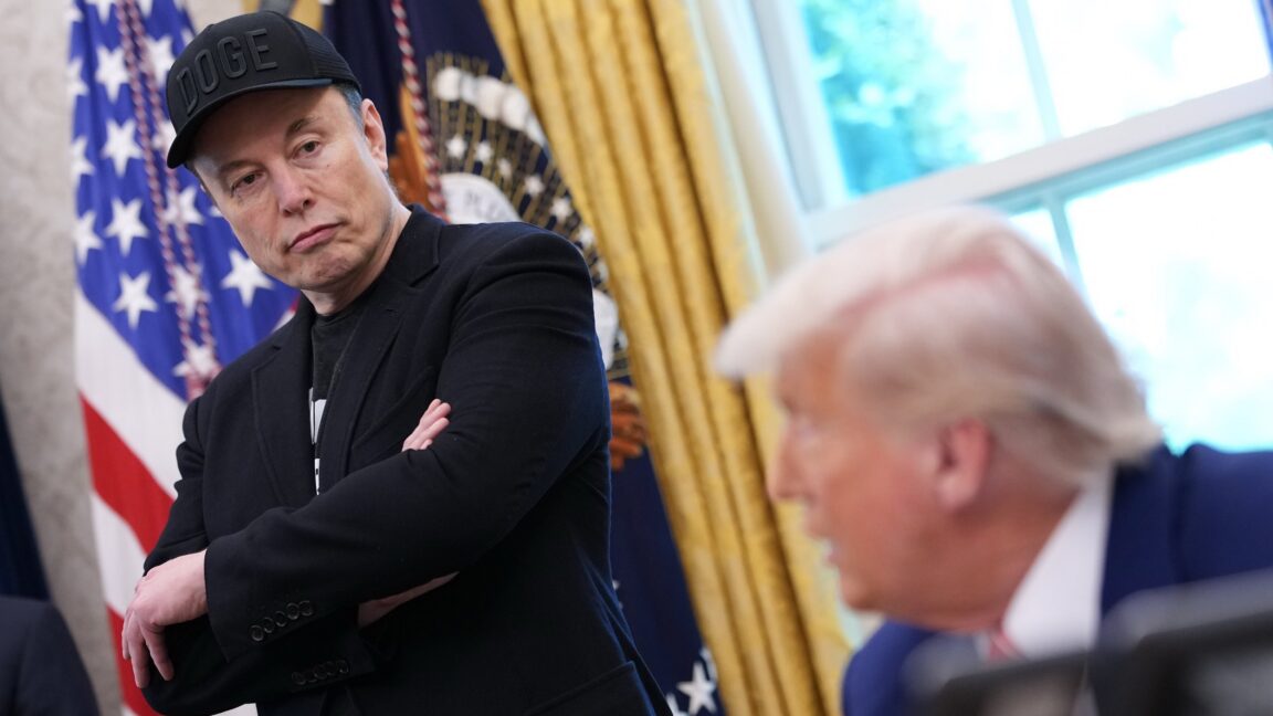 Elon Musk stands with his arms crossed, wearing a DOGE cap, in the Oval Office, near President Trump who sits at his desk while speaking.