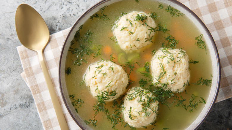 Top view of a bowl of matzo ball soup with a gold spoon and checkered napkin