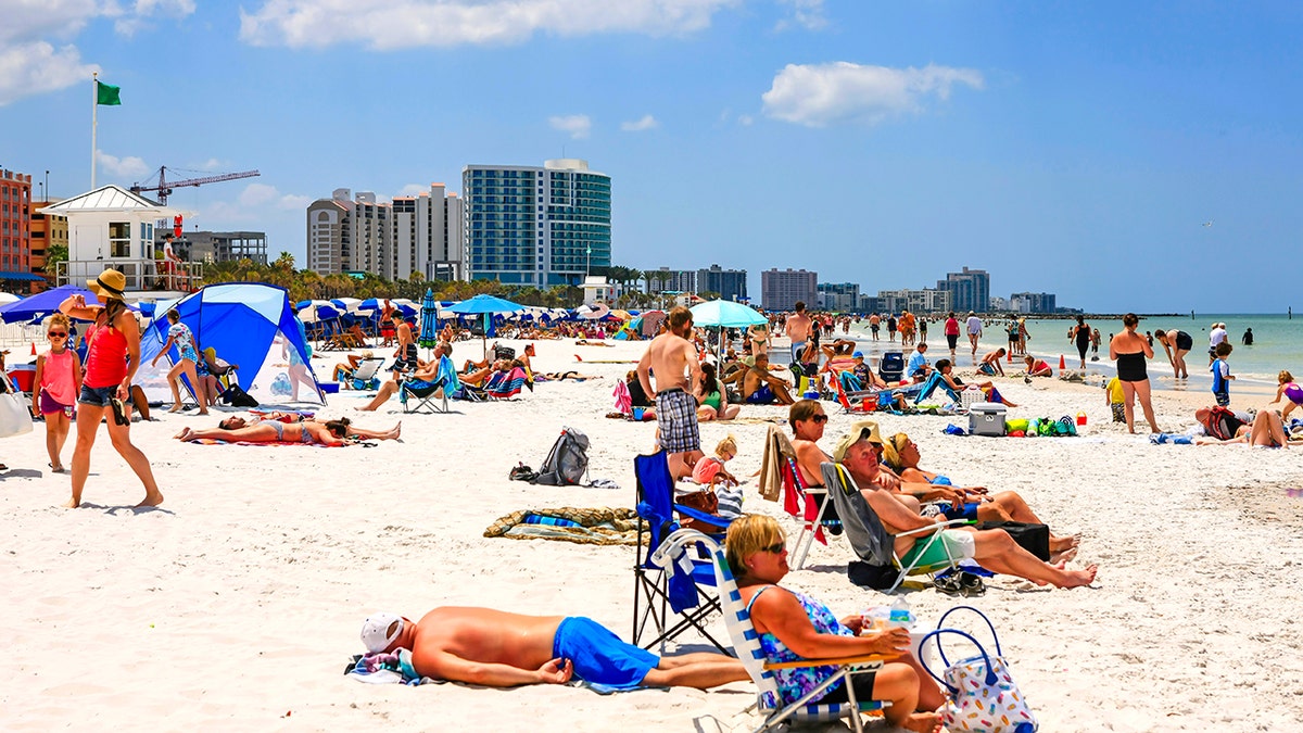 Crowded Florida beach with sunbathers, umbrellas, and beachfront high-rise hotels along the Gulf Coast.