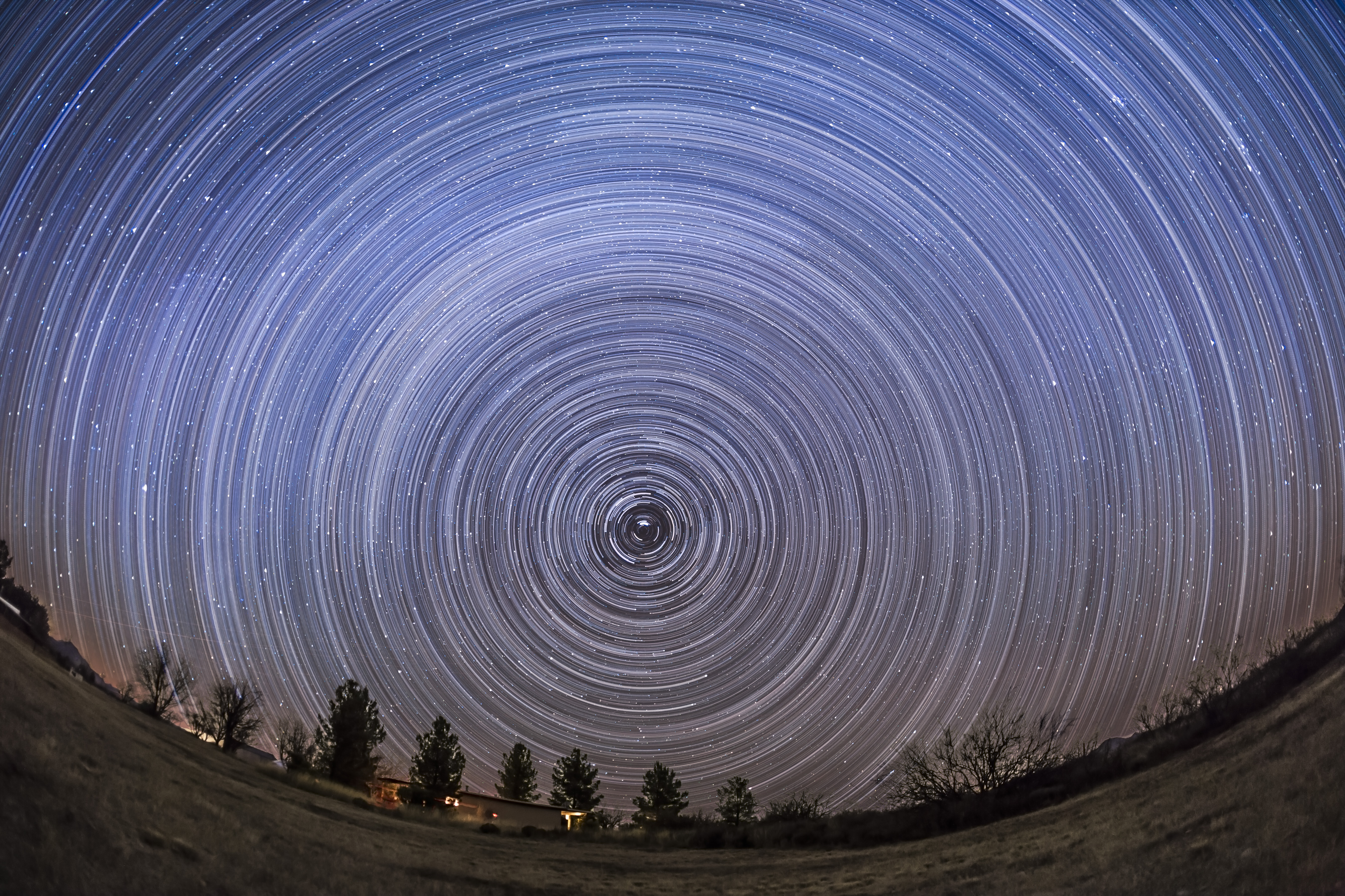 A timelapse photo of the night sky showing startrails circling the North Star Polaris above a field lined with trees.