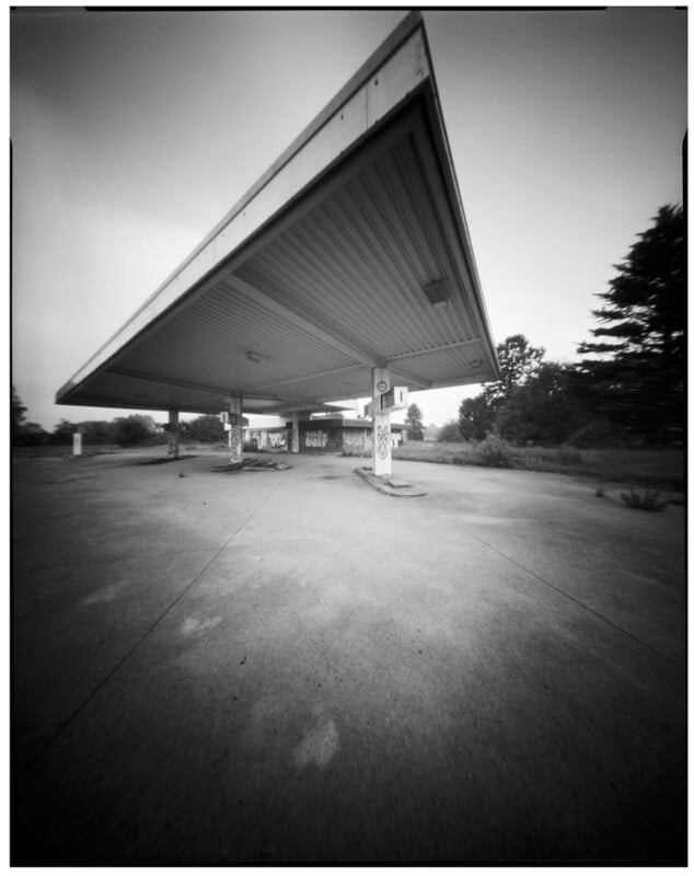 Wide-angle black and white photo of an abandoned gas station with graffiti on the columns, an empty forecourt, and overgrown vegetation in the background under a large, angular canopy.